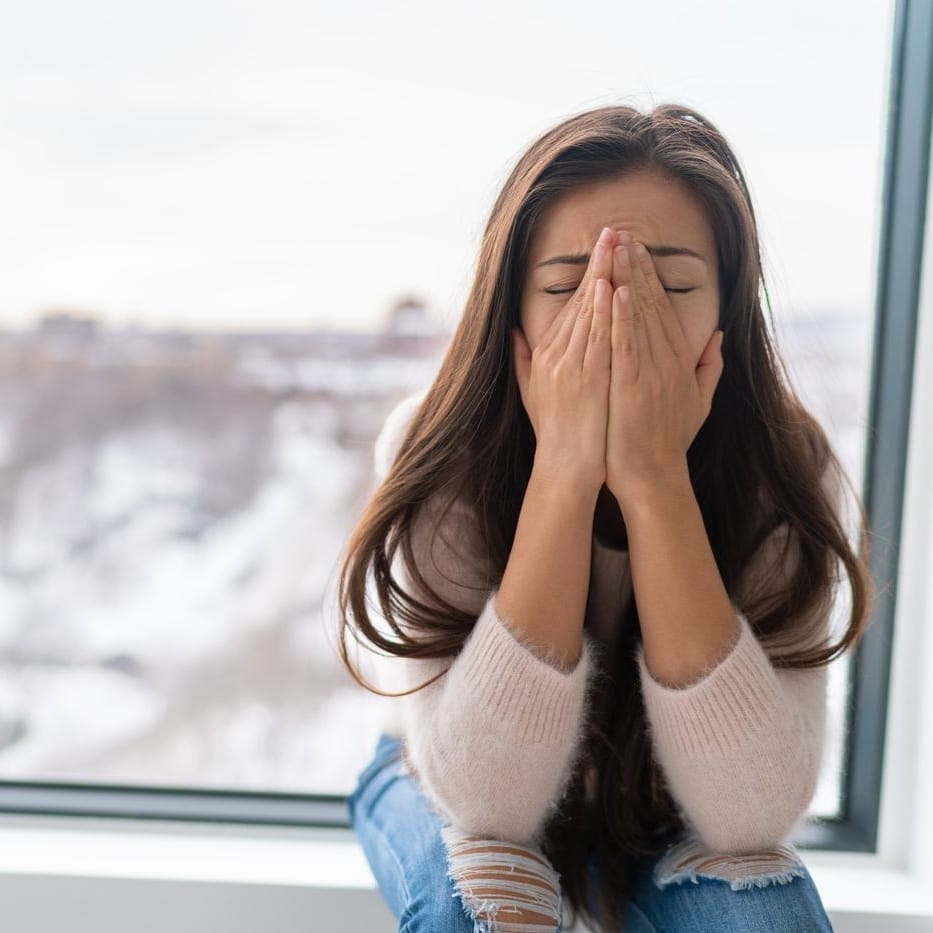 Woman sitting by a window with her face in her hands, reflecting the need for online anxiety counseling.