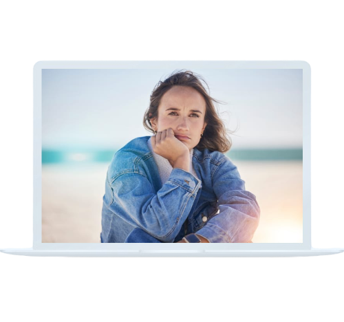 A woman sitting on a beach, looking thoughtful while featured on a laptop screen, showing accessibility through online drug addiction treatment.