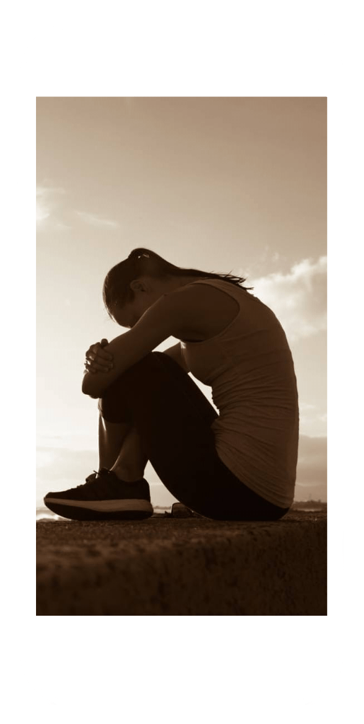 A woman sitting by the ocean at sunset with her head resting on her knees, conveying the struggle addressed through online trauma counseling.