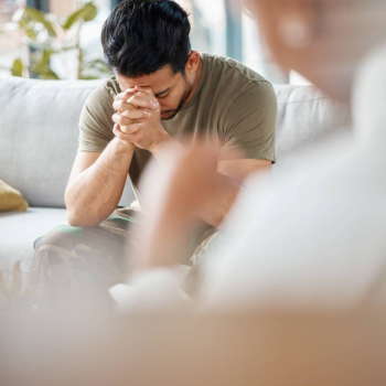 A man sitting on a couch with his head in his hands during a counseling session, showing the weight of PTSD and the support of online trauma counseling.