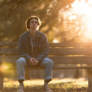 A young man sits on a wooden bench at sunset, reflecting quietly, symbolizing support found in online OCD counseling.